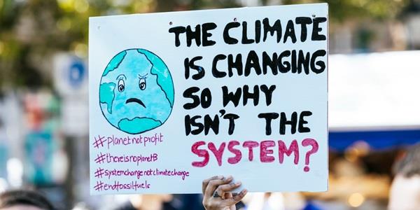 Someone holding a sign that shows a drawing of the earth and the text THE CLIMATE IS CHANGING SO WHY ISN'T THE SYSTEM?