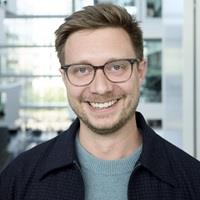 Photo of Tim. He is smiling, wears glasses, and the ITU building is visible in the background.