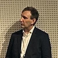 heashot of a white man with black jacket and white shirt looking to the right and not straight in the camera lens. The background is brown and dotted.