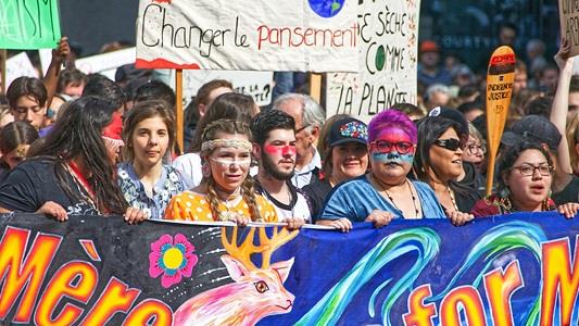 Crowd of indigenuos people behind large colourful banners in a climate protest. One of the signs held ud reads 'change le pansement'